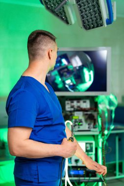 Cerrah parlak ameliyatları izler. Mavi önlüklü bir cerrah cerrahi görüntüler gösteren bir monitör üzerinde yoğunlaşır, hassas bir ameliyatı denetler..