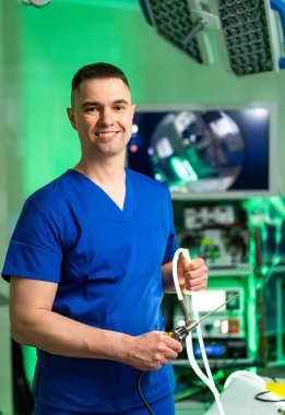 Cerrah cerrahi müdahaleye hazırlanıyor. Mavi önlüklü bir erkek cerrah gülümseyerek modern ve iyi donanımlı bir ameliyathanede aletleri tutuyor..