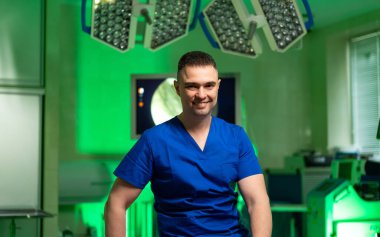 Ameliyathanede gülümseyen bir cerrah. Mavi önlük giyen bir cerrah, modern bir ameliyathanede, parlak cerrahi ışıklarla donatılmış, kendinden emin bir şekilde gülümser..