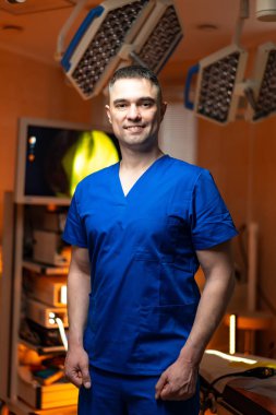 Mavi önlüklü bir cerrah. Bir cerrah, iyi aydınlatılmış bir tıbbi tesiste, ameliyata hazır mavi önlüklerle güvenle durur..