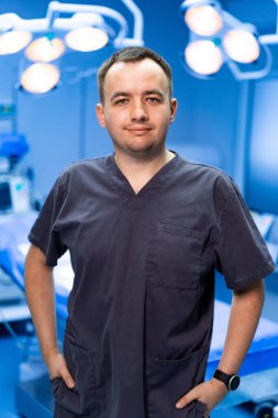 Cerrah emin bir şekilde ameliyathanede poz veriyor. Bir cerrah elleri kalçasında durur, tıbbi aletlerle donatılmış iyi aydınlatılmış bir ameliyathanede gülümser..