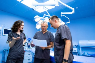 Ameliyat ekibi birlikte çalışır. Bir grup doktor hasta detaylarını iyi donanımlı bir ameliyathanede ameliyata hazırlanırken tartışıyor..