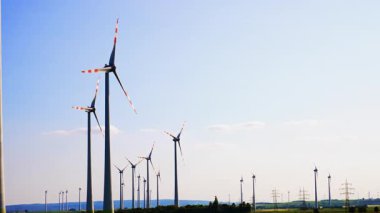 Türbinler temiz enerji üretiyor. Rüzgâr türbinleri açık mavi gökyüzüne karşı dönerek kırsal bir alanda yenilenebilir enerji üretimi sergiliyor..