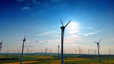 Mavi bulutların altında türbinler. Bereketli yeşil bir arazide, dağınık bulutlu mavi bir gökyüzünün altında sayısız rüzgar türbini dimdik ayakta duruyor..