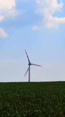 Mavi gökyüzüne karşı uzun türbin. Bir rüzgar türbini, parçalı bulutlu bir gökyüzünün altında yemyeşil alanlar üzerinde yavaşça döner. Yenilenebilir enerji devrede.