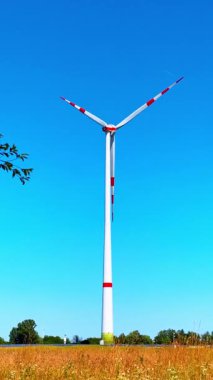 Uzun rüzgar türbini açık mavi gökyüzüne karşı. Uzun bir rüzgar türbini, berrak mavi gökyüzünün altında canlı bir manzarayla çevrili bir tarlada belirgin bir şekilde duruyor..