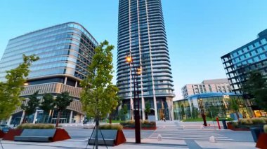 Şehir meydanı modern tasarım. Yüksek binalar, günbatımında çağdaş kentsel tasarımı yansıtan yeşillik ve banklarla dolu canlı bir plazanın çerçevesini oluşturuyor..