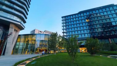 Şehir modern mimarisi. Yeşil bir alanı çevreleyen cam cepheli binalar, hareketli bir şehir bölgesinde akşam ışıklarıyla aydınlatılıyor..