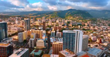 Honolulu, Hawaii, ABD 'nin modern şehir manzarası üzerinde uçuş. Arkaplanda koyu bulutların altında, yamaçlarda evleri olan yeşil dağlar..