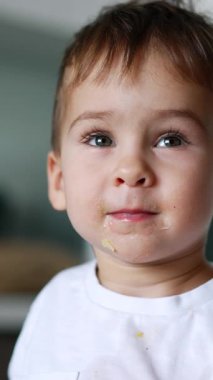 Yüzünde yemek olan sevimli bir oğlan çocuğu mutfakta oturuyor. Şirin çocuk kaşığını tabağına vuruyor. Kapatın. Dikey video