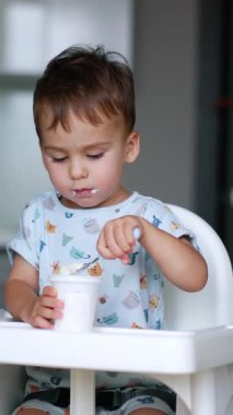 Güzel bir çocuk mutfakta yüksek bir sandalyede oturuyor. Kafkasyalı erkek bebek bir fincandan yoğurt yiyor. Dikey video