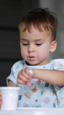 Güzel beyaz bebek mutfakta oturup fincandan yoğurt yiyor. Bebek sağlıklı bir şeyler yiyor. Dikey video