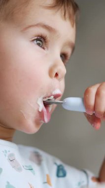 Tatlı küçük çocuk ağzına bir kaşık süt alıyor. Yoğurt yiyen çocuğun düşük açılı görüntüsü. Dikey video