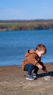Nehir kıyısında yürüyen 18 aylık bir çocuk. Küçük çocuk taşları alır ve suya atar. Dikey video