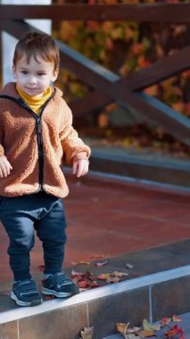 Kahverengi ceketli şirin erkek bebek verandada duruyor. Güler yüzlü çocuk merdivenden mutlu bir şekilde atlıyor. Dikey video