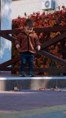 Kahverengi ceketli sevimli çocuk verandada. Şirin oğlan kuru yaprağı alıp öne fırlatıyor. Dikey video