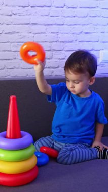 Mavi tişört giyen, kanepede oyuncakla oynayan küçük sakin bir çocuk. Piramidi birleştiren tatlı bebek. Dikey video