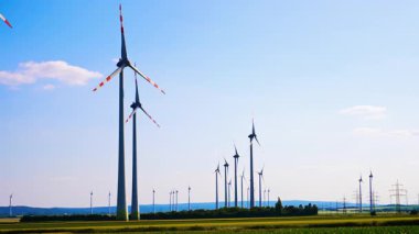 Türbinler temiz enerji üretir. Rüzgar türbinleri, açık mavi bir gökyüzü altında gündüz saatlerinde rüzgar enerjisini kullanan yeşil bir arazide dimdik dururlar..