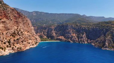 Yamaçlarda bitki örtüsü zayıf olan muhteşem kayalar. Denizin masmavi sularında hareket eden iki tekne..