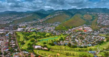 Ağaçların büyüdüğü güzel yeşil çayırlar golf oynamak için kullanılıyor. Honolulu, Hawaii, ABD 'deki dağların yamaçlarına ve vadiye dağılmış geniş yerleşim alanları. Gökyüzünde dramatik bulut manzarası var. Hava görünümü.