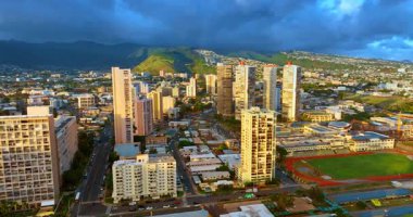 Gökdelenlerin tepeleri batan güneşin son ışınlarıyla aydınlanır. Gün batımında Honolulu manzarası. Arkaplandaki yeşil dağları dramatik bulutlar kaplıyor.