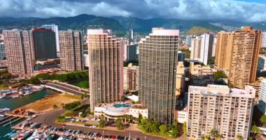 Pasifik Okyanusu kıyısındaki lüks, modern, yüksek katlı oteller. Waikiki kıyısı boyunca yat kulübünün manzaralı İHA uçuşu. Arka planda dağların üzerinde dramatik bulutlar var. Honolulu, Hawaii, ABD.