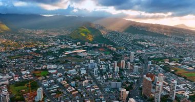 Modern Honolulu 'nun çeşitli binaları vadiye ve dağ yamaçlarına dağıldı. Batan güneşin ışınları yeşil dağı aydınlatan dramatik bulutların arasından geçer. Hava görünümü.