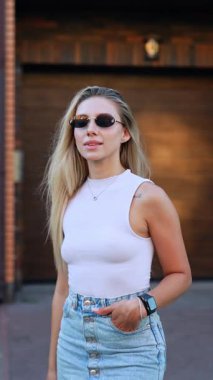 Sarışın, beyaz gözlüklü, beyaz bluzlu ve kot etekli kız dışarıda bekliyor. Güzel manken kameranın önünde poz vererek uzun saçlarına dokunur. Dikey video.