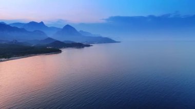 Pembe ışık, gün batımında deniz manzarasını boyuyor. Arkaplanda sisli dağların siluetleri.