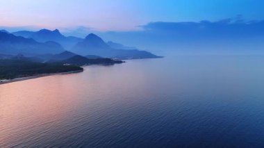 Pembe ışık, gün batımında deniz manzarasını boyuyor. Arkaplanda sisli dağların siluetleri.