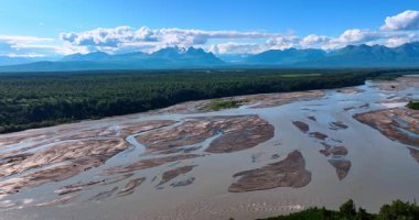Alaska 'nın göz kamaştırıcı manzarası. Sığ nehir her zaman yeşil olan ormandan akar. Pofuduk dağlar arka planda dağ sırasının üzerinde asılı. Hava görünümü.