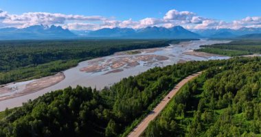 Büyüleyici yeşil ormanlarda akan sığ nehre yaklaşıyoruz. Arabalar ormanda nehir boyunca otoyola gider. Arkaplanda sıradağlar var. Alaska hava manzarası.