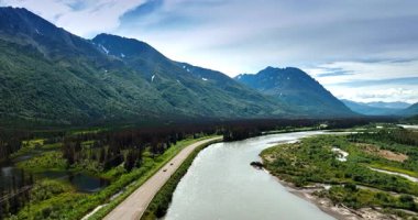 Yazın Alaska 'nın şaşırtıcı doğası. Dağın eteğinden akan nehrin üzerinden insansız hava aracı uçuşu. Arabalar nehir boyunca yol boyunca gider..