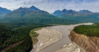 Bir nehrin kurumuş deltasının üzerinde uçuyor. Evergreen ormanları çevredeki toprakları kaplar. Arka planda bulutlu gökyüzünün altındaki yemyeşil dağlar. Alaska.