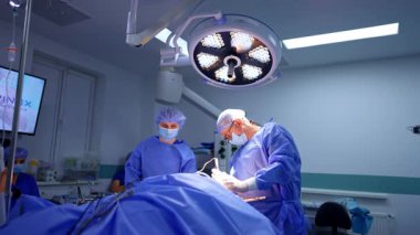 Hastane cerrahisinde cerrahlar. Mavi önlüklü iki cerrah modern bir ameliyathanede parlak ışıklar altında cerrahi bir operasyona odaklanıyor..