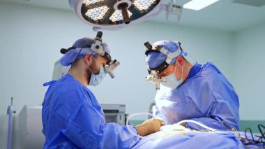 Cerrahlar ameliyathanede. Mavi önlüklü iki cerrah parlak bir ameliyathanede özel aletlerle ameliyata odaklanıyor..