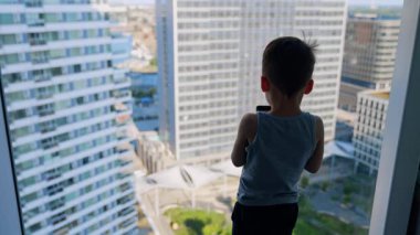 Çocuk otelden ufuk çizgisine bakıyor. Genç bir çocuk pencerenin kenarında durup, gün boyunca yüksek binalarla dolu canlı bir şehir siluetine bakıyor..