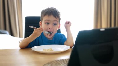 Çocuk yemek yiyor, televizyon seyrediyor. Genç bir çocuk yemek masasında makarna yerken tabletten eğlenceli bir şeyler izliyor..