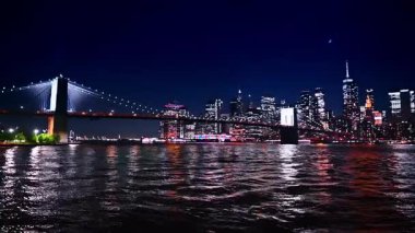 Nehrin karanlık suları metropolün birçok şehir ışığını yansıtır. Brooklyn Köprüsü ve New York gökdeleninin alçak açılı görüntüsü.