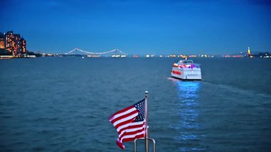Teknenin ön tarafında Amerikan bayrağı dalgalanır. Büyük beyaz nehir teknesi nehrin önünden geçiyor. Arka planda New York 'un parlak silueti.