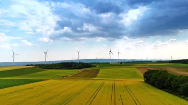 Yaz günü kırsaldaki çimenli çayırların üzerinden uçacağız. Arka planda birden fazla rüzgar türbini dönüyor. Yeşil enerji kavramı.
