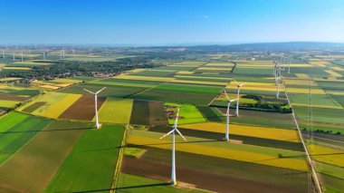 Türbinler temiz enerji üretir. Rüzgâr türbinleri açık mavi gökyüzünün altında yeşil alanlarda dönüyor, kırsal alanda yenilenebilir enerjiyi vurguluyorlar..