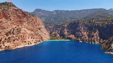 Nefes kesici uçurumlar ve sakin sular. Uçurumlar, sakin bir körfezi, mavi sularda parlayan güneş ışığını ve arka planda yeşil tepeleri kuşatıyor..