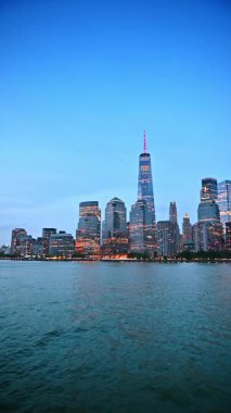 New York, ABD 'nin ufuk çizgisinde ışıkları yanan çarpıcı modern binalar. Su yüzeyinden yüksek binaların alçak açılı görüntüsü. Dikey video.