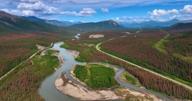 Alaska 'nın geniş vadisindeki dallanan nehrin üzerindeki görüntüler. Arabalar otobanın yanından geçerek arka plandaki dağlara doğru gidiyor..