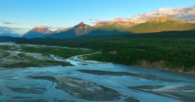 Güzel vadide akan sığ nehir. Batan güneş, dağların tepelerini kısmen pembe bulutlarla aydınlatıyor. Alaska vahşi doğa.
