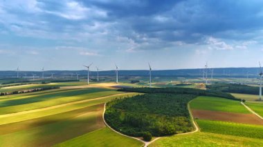 Rüzgar türbinleri yeşil enerji üretir. Rüzgâr türbinleri, dalgalı tarlaların ve çarpıcı bir gökyüzünün altında canlı yeşil bir ormanın üzerinde dimdik durur..