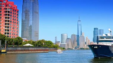 Küçük beyaz tekne yeşil rıhtım boyunca kameraya yaklaşıyor. Çapanın yanında büyük bir tekne duruyor. Arkaplanda Manhattan 'ın silüeti.