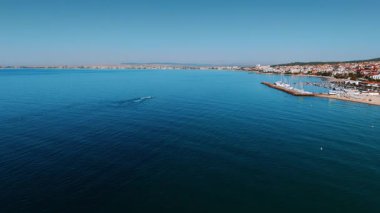 Canlı bir sahil kasabasının kıyı manzarası. Parlak güneşli bir günde sakin bir deniz ve kıyı kasabasında hareketli bir plaj sergileniyor..