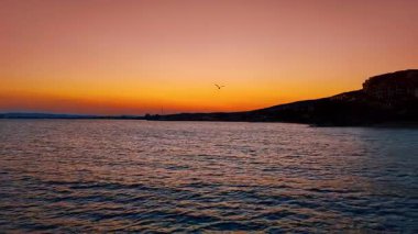 Sakin deniz sularının üzerinde güzel bir gün batımı. Canlı turuncu ve mor gökyüzü alacakaranlıkta sakin okyanus sularına yansıyarak kıyıya yakın sakin bir atmosfer yaratır..